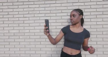 Bu yavaş çekim videosu, şık siyah bir kadının, akıllı telefonuyla dokulu beyaz tuğla bir arka planda özgüven selfie çekerken görüntülerini içeriyor. Onun odaklanmış ifadesi