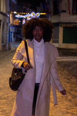 Bu resim, şehir ışıklarının parlaklığı ve şenlikli dekorasyonlarla aydınlatılmış, gece kaldırım taşı bir sokakta yürüyüş yapan şık bir kadını tasvir ediyor. Kendine güveni ve doğrudan bakışları