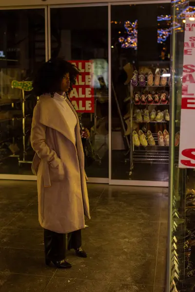 stock image The warmth of a city evening is encapsulated in this image, where a woman stands by a shop window, the word SALE mirrored in bold letters. The festive lights twinkle in the background, suggesting a