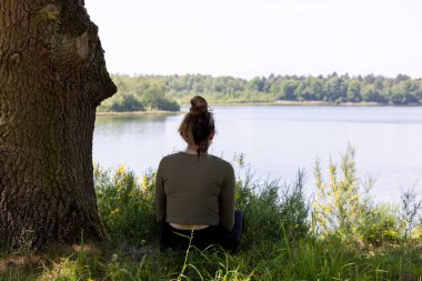 This evocative image depicts a woman from behind, sitting in stillness by a tranquil lake. Flanked by a robust tree trunk and overlooking calm waters, her posture suggests deep relaxation or clipart