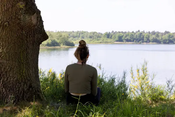 stock image This evocative image depicts a woman from behind, sitting in stillness by a tranquil lake. Flanked by a robust tree trunk and overlooking calm waters, her posture suggests deep relaxation or