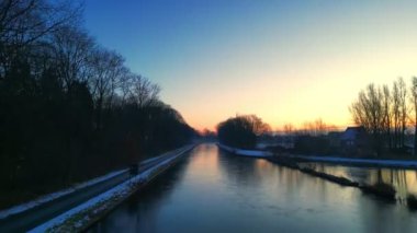 Bu sakin stok görüntüsü, donmuş bir kanalın gün doğumunun yükselen renklerini yansıttığı sakin bir kış sabahının güzelliğini yakalar. Kamera perspektifi su yüzeyine yakın ve alçak.