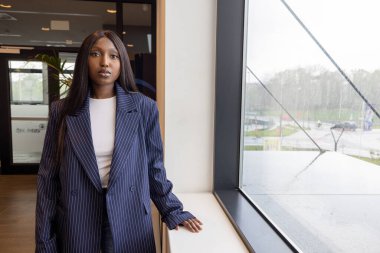 Bu görüntü, çizgili takım elbise giymiş, dışarıda yağmurlu bir gün manzarası olan büyük bir pencerenin yanında duran genç bir Afro-Amerikalı kadını profesyonel bir ortamda yakalıyor. Onun sakin tavrı.