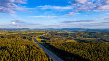 This aerial photograph captures the sweeping view of the E42 highway as it cuts through the verdant landscape of the Hautes Fagnes area near Emmels. The lush green forests dominate the scene, with clipart