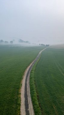 Bu hava görüntüsü, sisli bir sabahta canlı yeşil tarlaları kesen dar bir köy yolunu yakalıyor. Yol, sislerin arasından göz alıcı bir görüntü çizgisi oluşturur.