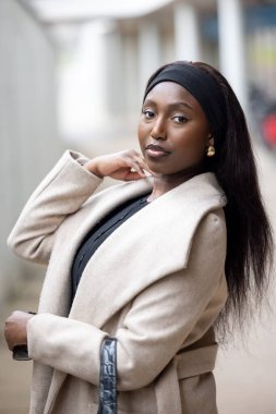 This portrait captures a young Black woman striking a confident pose on a city street, dressed in a stylish beige coat over a black ensemble. Her relaxed posture, combined with a thoughtful expression
