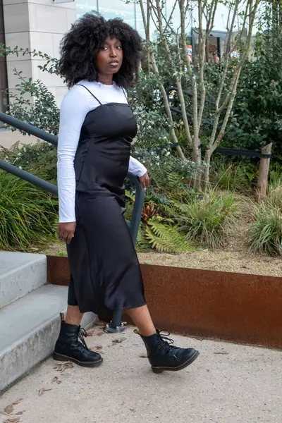 stock image This image features a Black woman descending outdoor stairs in an urban setting, dressed in a stylish black dress layered over a long-sleeve white top, paired with black combat boots. Her natural