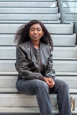 A woman wearing a leather jacket is seated on a staircase, exuding a sense of comfort and confidence. She looks relaxed and happy in the setting