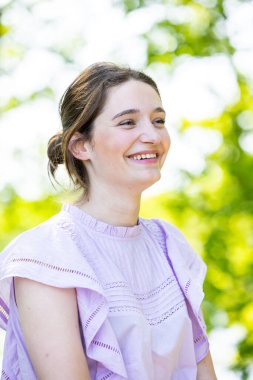 Bu görüntü, ince bir gülümsemesi olan, güneşli bir bahçede bir anın tadını çıkaran genç bir kadının yan profilini gösteriyor. Güneş ışığı yaprakların arasından süzülüyor, yüzüne ve yüzüne yumuşak bir parıltı saçıyor.