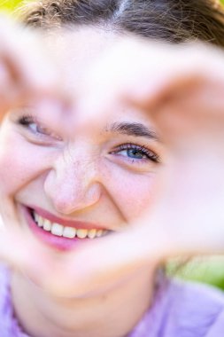 This is a playful and intimate close-up of a young woman seen through a heart-shaped hand gesture shes making, which frames her sparkling eyes and cheerful smile. The focus is on her eyes and the clipart