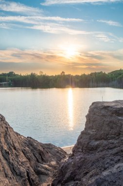 The sunset creates a stunning scene on a calm lake, with rocks in the front and a peaceful forest in the back