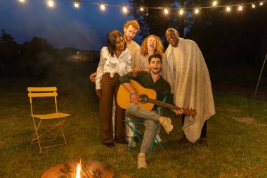 Arkadaşların eğlenceli bir akşamın tadını çıkarıyorlar, kamp ateşi altında gitar çalıyorlar ve neşeli bir ortam yaratıyorlar.