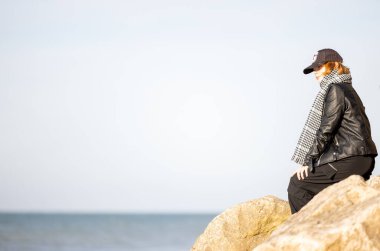 Sahilde kayalara oturmuş deri ceket, kep ve eşarp giymiş bir kadın manzaranın tadını çıkarıyor.