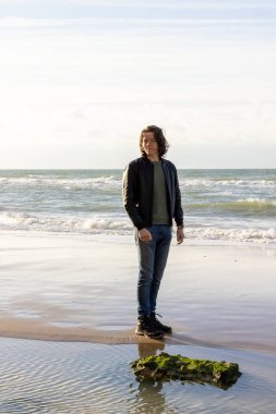 Günlük giysiler içinde bir adam sahilde duruyor, okyanusa doğru gülümsüyor, manzaralı deniz manzarasının ortasında neşe saçıyor.