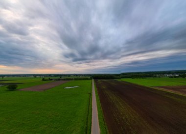 Sakin bir kır manzarası sergileniyor. Sessiz bir yol ile bereketli arazide dramatik bulutlu gökyüzünün altında dönüyor.