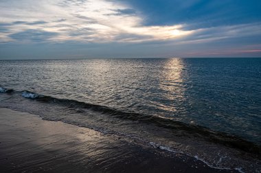 Bu görüntü plajda gün batımının sakin bir tasviri, güneşin yansıması okyanus dalgaları üzerinde parıldıyor. Gökyüzü, yumuşak gri ve mavilerin karışımı ve biraz ılık portakal.