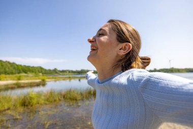 A woman in a blue sweater joyfully looks at the calm lakeside scenery, immersed in natures tranquility clipart