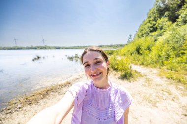 Güneşli bir günde göl kenarında yemyeşil ve açık gökyüzüyle çevrili bir kadın mutlu bir selfie çekiyor.