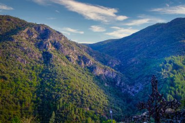 Bereketli yeşillik ve görkemli dağlarla çevrili çarpıcı bir vadi, berrak mavi gökyüzünün altında, kabarık bulutlarla.