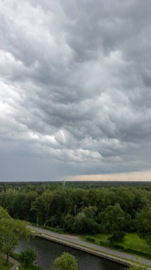 Sakin bir manzara, bir orman, kanal ve iz üzerinde asılı fırtına bulutları ile tasvir edilir, karamsar bir atmosfer yaratır