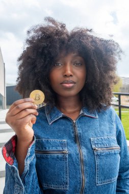 Kot ceketli, açık havada elinde bitcoin tutan, modern şehir ortamında kripto para yatırımı yapan kendine güvenen bir kadın.