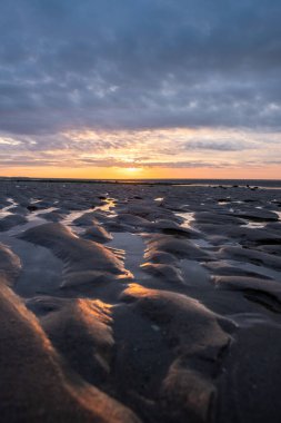 Kumda ve dramatik gökyüzünde canlı renklerle baş döndürücü bir günbatımı sahilde sakin bir atmosfer yaratıyor.