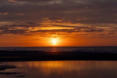 Huzurlu bir sahildeki sakin sularda yansımalarla ufku aydınlatan parlak bir altın gün batımı..