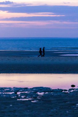 Alacakaranlıkta kumsalda gezinen bir çiftin silueti, Gökyüzünde mor ve mavi renklerin sakin bir karışımı altında
