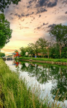 Etrafı yeşillik ve bulutlu gökyüzü ile çevrili sakin bir kanalda güzel yansımalarla donatılmış güneşli bir gün batımı..