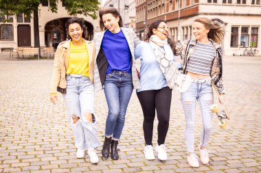 Güneşli bir günde, canlı, kentsel bir ortamda birlikte yürüyüp gülen dört arkadaş, birbirlerine eşlik ederek eğleniyorlar.