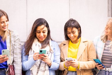 Çeşitli bir grup kadın açık havada akıllı telefonları kullanarak güneşli bir günde neşe ve dostluk gösteriyorlar.