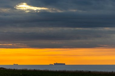 Uzak okyanus ufkunda bulutlarla dolu dramatik bir gökyüzünün ortasında nefes kesici bir gün batımı görünüyor.