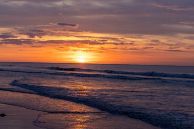 Okyanusun üzerinde huzurlu bir gün batımı, kıyıya vuran hafif dalgalar, kumsalda altın bir renk..