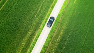 Dar bir kırsal yolda, yeşil yemyeşil tarlalarla çevrili, sakin bir çevrede siyah bir arabanın hava görüntüsü.