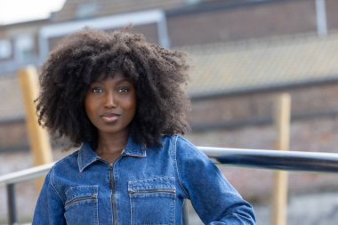 Afro saç stili olan kendine güvenen bir kadın, şehir arka planına dayanan kot ceketiyle dışarıda poz veriyor.