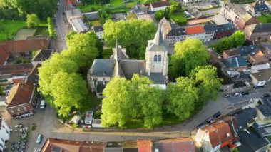 Sint-Pieters-Leeuw, Brüksel, Belçika, 4 Haziran 2024, görkemli küçük bir kasabadaki yemyeşil ağaçlarla çevrili tarihi bir kilisenin çarpıcı hava manzarası..