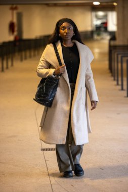 A woman walks confidently indoors in a chic beige coat, carrying a black handbag and wearing casual attire. clipart