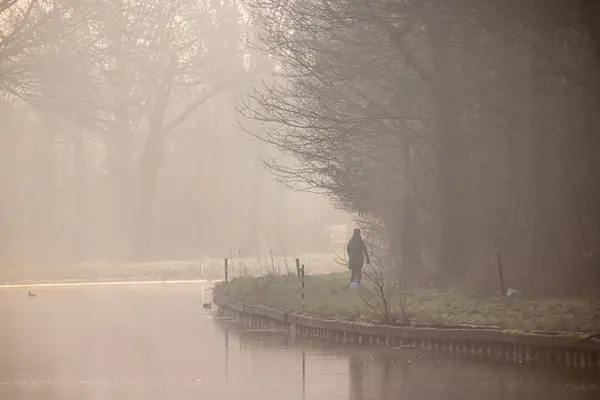 stock image A lone person walking by a foggy riverbank, surrounded by trees, creating a tranquil and peaceful scene