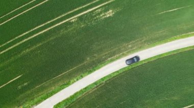 Siyah bir araba, virajlı bir yoldaki yemyeşil tarım tarlalarından geçiyor, İHA görüntüleriyle çekiliyor.