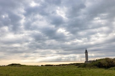 A picturesque lighthouse stands tall under a dramatic cloudy sky with open grass fields in the countryside. clipart