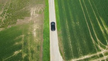 Siyah bir araba, yeşil ve kahverengi tarlalarla çevrili kırsal bir yolda ilerliyor.