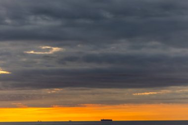 Okyanusun üzerinde kara bulutlar, ufuktaki gemileri aydınlatan canlı bir günbatımı dramatik bir deniz manzarası yaratır.