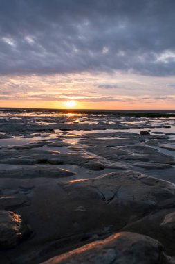 Şok edici bir günbatımı başımızın üstünde dramatik bulutlarla alçak gelgit sırasında kayalık bir kıyı manzarasını aydınlatıyor.