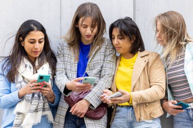 Açık havada akıllı telefonlar kullanarak modern bağlantıyı ve etkileşimi temsil eden günlük kıyafetli dört genç kadın.