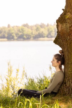 Genç bir kadın gölün kenarındaki bir ağaçta oturur, güneşli bir günde huzur içinde, etrafı sükunetle çevrili.