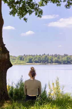 Kadın gölün kenarında oturuyor, sakin doğanın ve sükunetin tadını çıkarıyor, bir ağacın altında, sakin bir şekilde..