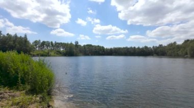 Yoğun ağaçlar ve yeşilliklerle çevrili dingin bir göl açık bir günde, kabarık bulutlarla, resim gibi bir manzara yaratıyor.