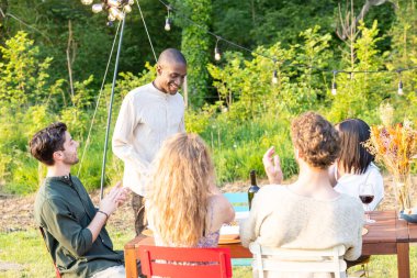 Bir grup arkadaş, mum ışığıyla aydınlatılmış yemyeşil bir bahçede keyifli bir akşam yemeği yiyor.