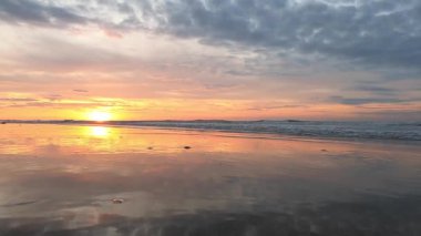 Sakin sahil üzerinde renkli gökyüzü ile sakin gün batımı, yansıtan bulutlar ve dalgalı suda parlayan güneş.