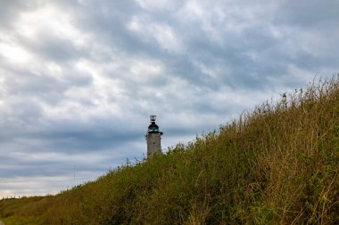 Arka planda dramatik bulutlar, çimenli bir tepenin üzerinde duran bir deniz feneri manzaralı bir kıyı manzarası sunuyor.